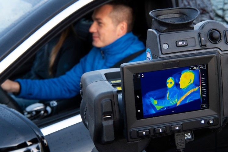 Man in blue uniform inspects car safety features with thermal imaging camera.