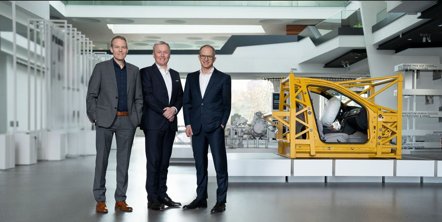 Three men in suits standing together in front of a yellow automotive safety frame with an airbag.