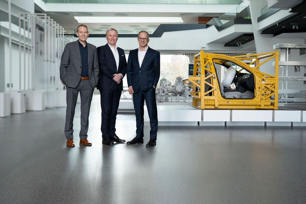 Three men in suits standing together in front of a vehicle safety testing frame.
